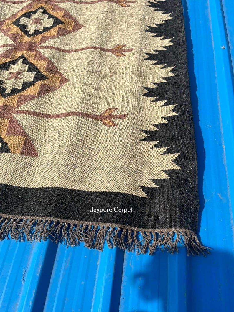 May include: A beige and black woven rug with a geometric pattern. The rug features diamond shapes in brown, black, and tan, with a black border and fringe. The text "Jaypore Carpet" is visible.
