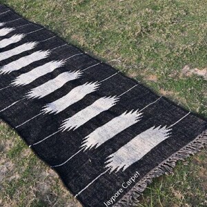 May include: A black and white rug with a repeating pattern of white, stylized brushstrokes. The rug has a fringed edge and is lying on green grass. The text "Jaypore Carpet" is visible on the rug.
