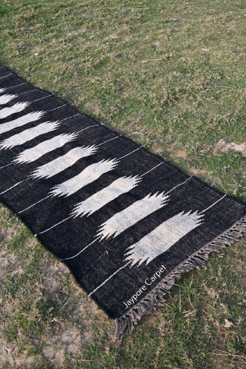 May include: A black and white rug with a repeating pattern of white, stylized brushstrokes. The rug has a fringed edge and is lying on green grass. The text "Jaypore Carpet" is visible on the rug.