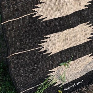 May include: Black and cream woven rug with a geometric design. The rug has bold black stripes with jagged, cream-colored accents. It has a fringed edge and the text "Jaypore Carpet" is visible.