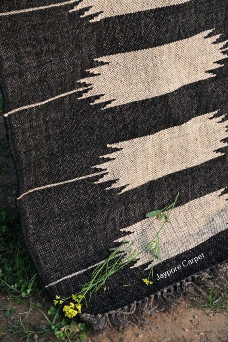 May include: Black and cream woven rug with a geometric design. The rug has bold black stripes with jagged, cream-colored accents. It has a fringed edge and the text "Jaypore Carpet" is visible.