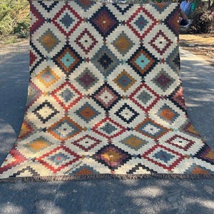 May include: A large, rectangular rug with a geometric diamond pattern in shades of red, blue, yellow, and brown. The rug has a cream-colored background and a fringed edge. The text "Jaypore Carpet" is visible on the bottom left.