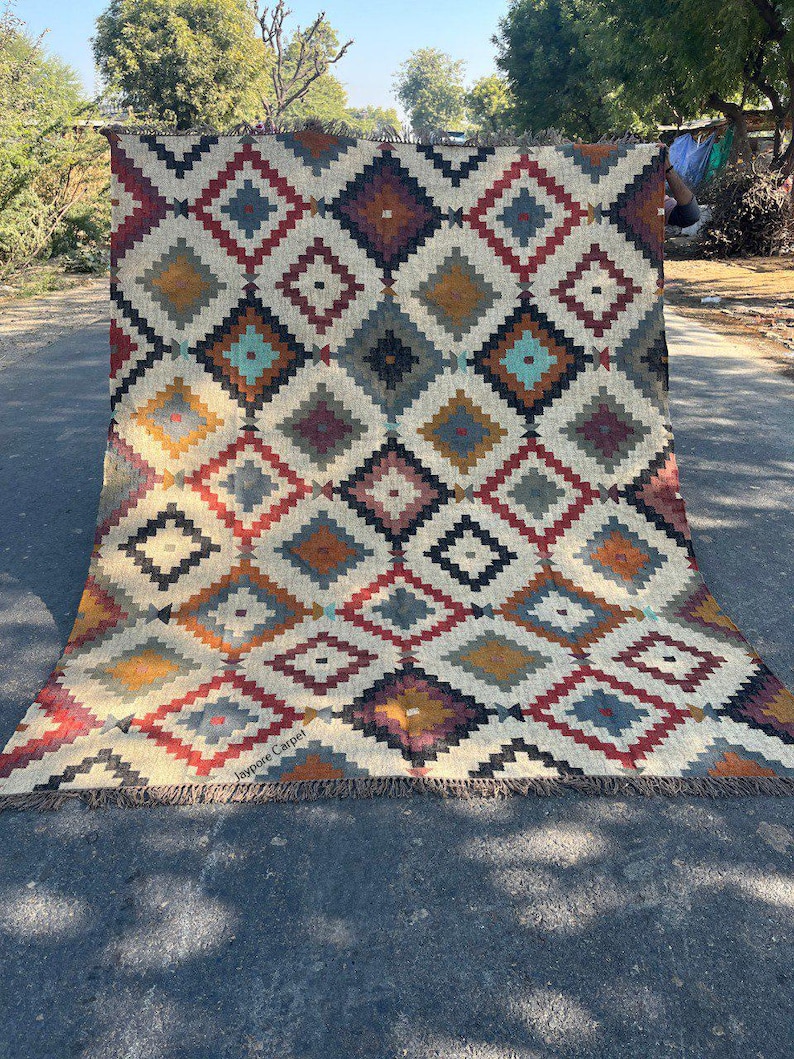 May include: A large, rectangular rug with a geometric diamond pattern in shades of red, blue, yellow, and brown. The rug has a cream-colored background and a fringed edge. The text "Jaypore Carpet" is visible on the bottom left.