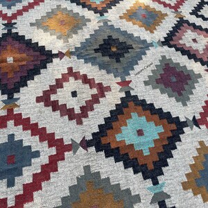 May include: A close-up of a woven rug with a geometric pattern. The rug features diamond shapes in various colors, including red, blue, brown, and teal, set against a light gray background. The design has a tribal or Southwestern aesthetic.