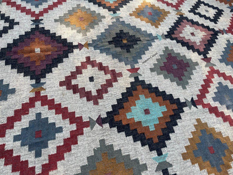 May include: A close-up of a woven rug with a geometric pattern. The rug features diamond shapes in various colors, including red, blue, brown, and teal, set against a light gray background. The design has a tribal or Southwestern aesthetic.