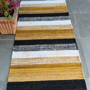 May include: A rectangular rug with alternating horizontal stripes of black, white, and gold. The rug is made of woven material and has a textured appearance. The rug is placed on a gray tiled surface.