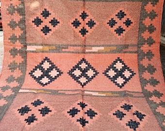 May include: A peach-colored woven rug with a geometric pattern. The design features repeating diamond shapes in dark blue and white, framed by a gray border. The rug has the text "Jaypore Carpet" at the bottom.