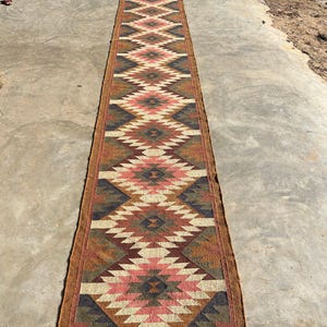 May include: A long, rectangular runner rug with a geometric diamond pattern in shades of brown, pink, cream, and navy blue. The rug has a fringed edge and is made of a woven material. The rug is approximately 10 feet long.