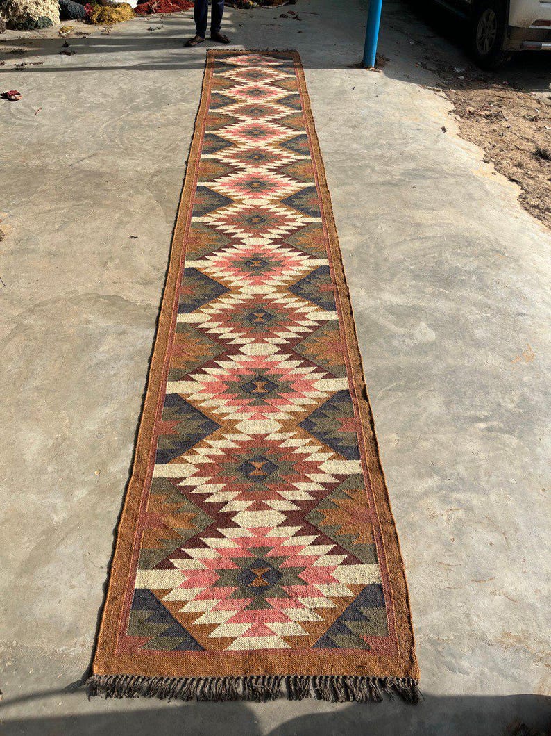 May include: A long, rectangular runner rug with a geometric diamond pattern in shades of brown, pink, cream, and navy blue. The rug has a fringed edge and is made of a woven material. The rug is approximately 10 feet long.