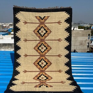 May include: Rectangular beige rug with a geometric pattern in brown and black. The rug features a central design of diamond shapes and arrow-like motifs, framed by a black border with a zigzag edge. The rug has a fringed edge and the text "Jaypore Carpet".