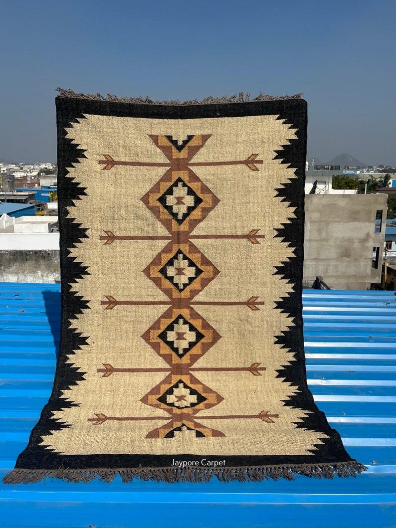 May include: Rectangular beige rug with a geometric pattern in brown and black. The rug features a central design of diamond shapes and arrow-like motifs, framed by a black border with a zigzag edge. The rug has a fringed edge and the text "Jaypore Carpet".