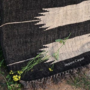 May include: A black and white woven rug with a geometric design. The rug has two large, light-coloured, horizontal shapes against a dark background. The text "Jaypore Carpet" is visible. Green plants and yellow flowers are scattered around the rug.