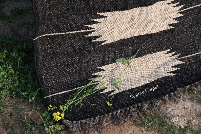 May include: A black and white woven rug with a geometric design. The rug has two large, light-coloured, horizontal shapes against a dark background. The text "Jaypore Carpet" is visible. Green plants and yellow flowers are scattered around the rug.