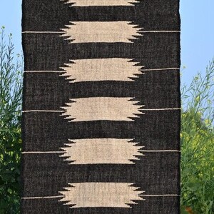 May include: Black and beige woven rug with a geometric pattern. The rug features horizontal bands of beige shapes against a black background. The bottom edge has a fringe. The text "Jaypore Carpet" is visible.