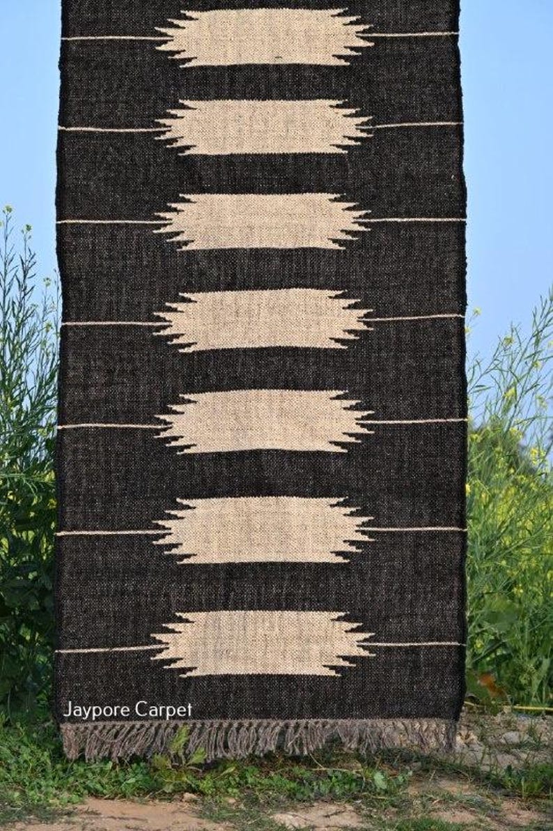 May include: Black and beige woven rug with a geometric pattern. The rug features horizontal bands of beige shapes against a black background. The bottom edge has a fringe. The text "Jaypore Carpet" is visible.