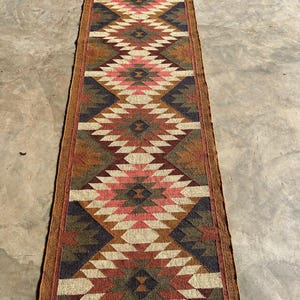 May include: A long, rectangular runner rug with a geometric diamond pattern. The rug features a color palette of red, pink, brown, beige, and navy blue. The rug is made of woven material and has a brown border.