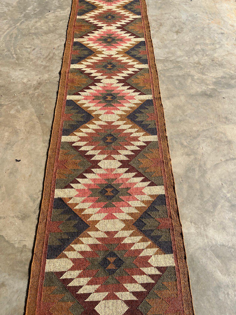 May include: A long, rectangular runner rug with a geometric diamond pattern. The rug features a color palette of red, pink, brown, beige, and navy blue. The rug is made of woven material and has a brown border.