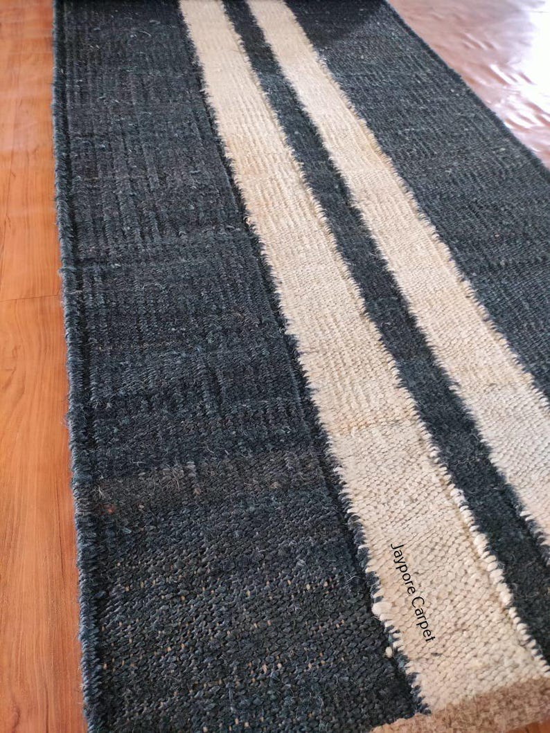 May include: A rectangular rug with a dark blue background and two off-white stripes running lengthwise. The rug has a textured appearance and a fringed edge. The text "Jaypore Carpet" is visible on the rug.