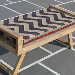May include: A wooden bench with a woven seat featuring a navy and beige zigzag pattern. The seat has a red stripe along the edge. The bench has a natural wood finish and angled legs, suitable for indoor or outdoor use.