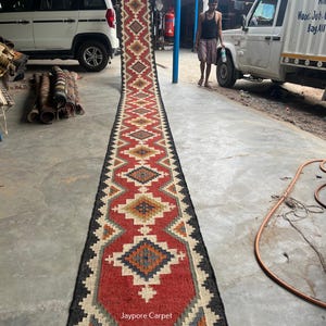 Peut inclure: Un long tapis de couloir étroit présentant un motif géométrique répétitif en losanges dans des tons de rouge, crème, bleu et marron, avec une bordure noire. Le texte 'Jaypore Carpet' est imprimé en blanc près du bas de la surface du tapis.