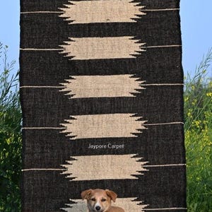 May include: A black and beige patterned rug with the text "Jaypore Carpet". The rug features a geometric design with horizontal beige shapes against a black backdrop. A small brown dog stands in front of the rug.