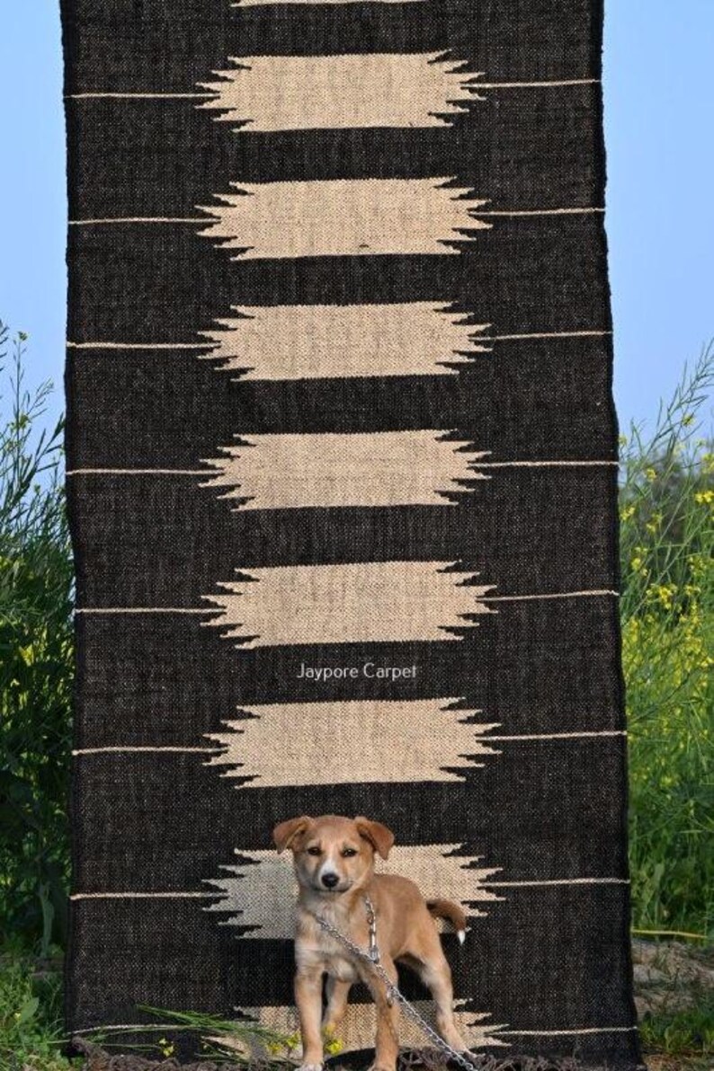 May include: A black and beige patterned rug with the text "Jaypore Carpet". The rug features a geometric design with horizontal beige shapes against a black backdrop. A small brown dog stands in front of the rug.