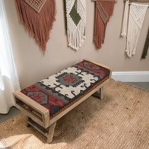 May include: A wooden bench with a rectangular seat featuring a colorful, patterned rug design in red, gray, and cream. The bench has a light wood frame and is placed on a woven jute rug. Decorative wall hangings are visible in the background.