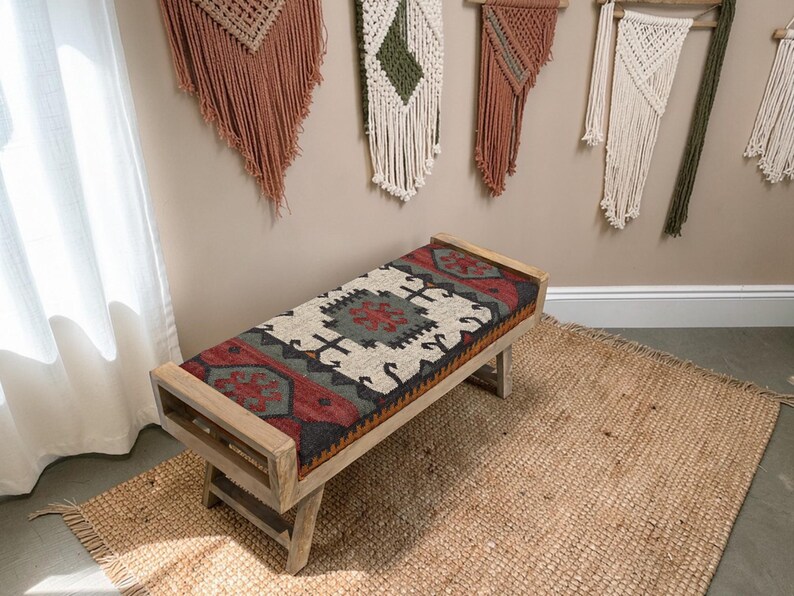 May include: A wooden bench with a rectangular seat featuring a colorful, patterned rug design in red, gray, and cream. The bench has a light wood frame and is placed on a woven jute rug. Decorative wall hangings are visible in the background.