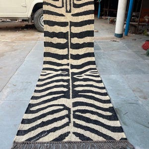 Puede incluir: Una alfombra rectangular con un patrón de rayas de cebra en negro y beige. La alfombra tiene un borde con flecos y el texto "Jaypore Carpet" es visible. La alfombra está sostenida por una persona.