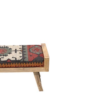 May include: A wooden bench with a rectangular seat featuring a patterned fabric in shades of red, green, black, and beige. The bench has a light-colored wooden frame and angled legs, suitable for indoor use.