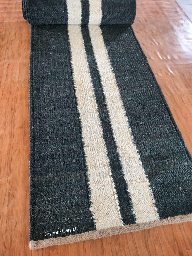 May include: A rolled-up, dark gray runner rug with two wide, cream-colored stripes running lengthwise. The rug is made of a textured material and is displayed on a wooden floor. The text "Jaypore Carpet" is visible on the bottom left corner.