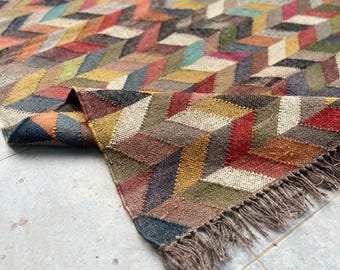 Pode incluir: Um tapete geométrico colorido com um padrão repetitivo de formas angulares em várias cores, incluindo vermelho, azul, amarelo e castanho. O tapete tem uma borda com franjas e está parcialmente dobrado.
