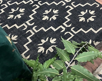 Puede incluir: Una alfombra negra y beige con un diseño geométrico. La alfombra presenta un patrón repetitivo de zigzags y formas florales. La alfombra está hecha de un material tejido y tiene un borde con flecos.