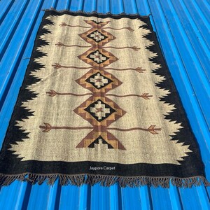May include: Rectangular beige rug with a geometric pattern in brown, black, and tan. The design features diamond shapes and arrow-like elements. The rug has a black border with fringe and the text "Jaypore Carpet".