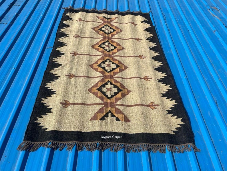 May include: Rectangular beige rug with a geometric pattern in brown, black, and tan. The design features diamond shapes and arrow-like elements. The rug has a black border with fringe and the text "Jaypore Carpet".