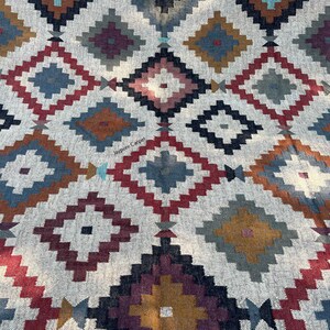 May include: A woven rug with a geometric diamond pattern in shades of red, blue, brown, and gray on a cream background. The rug features a repeating design of diamond shapes, creating a textured and visually appealing surface. The text "Jaypore Carpet" is visible.