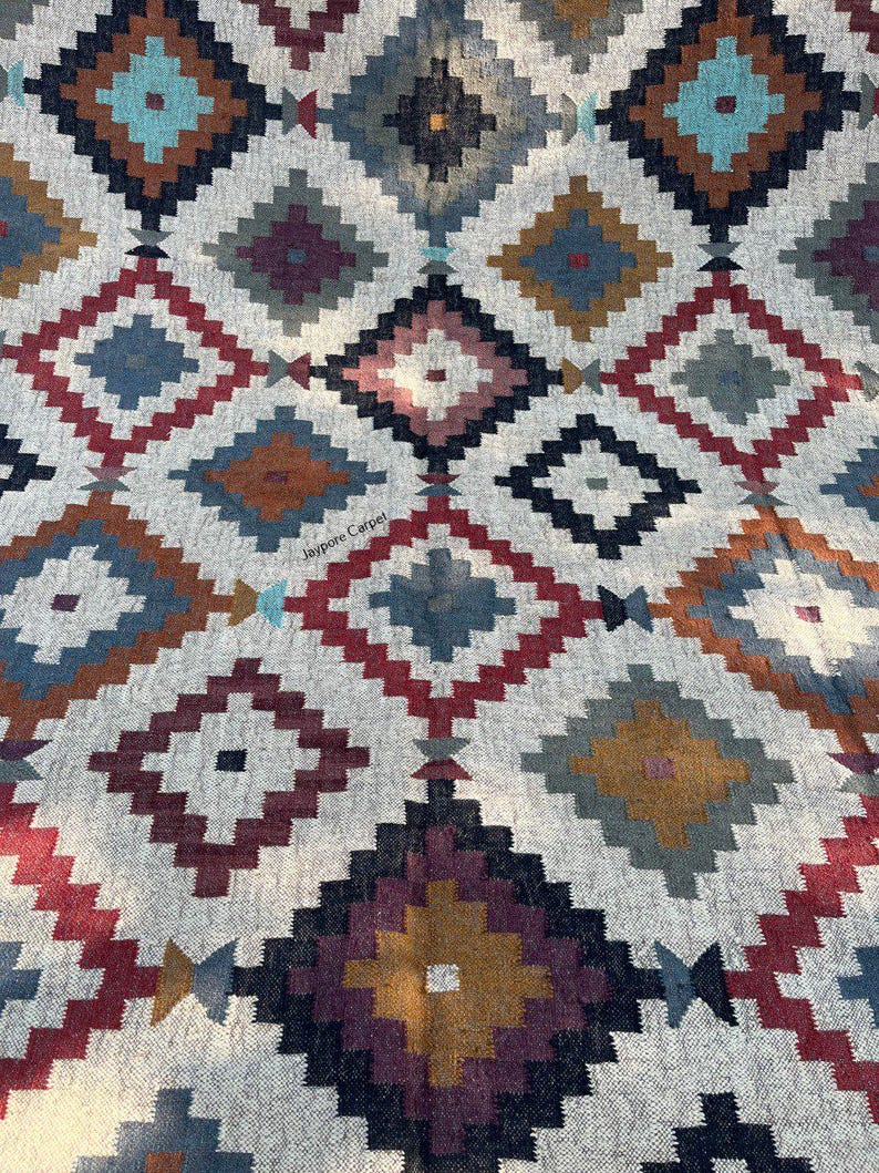 May include: A woven rug with a geometric diamond pattern in shades of red, blue, brown, and gray on a cream background. The rug features a repeating design of diamond shapes, creating a textured and visually appealing surface. The text "Jaypore Carpet" is visible.