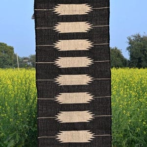 May include: A black and beige woven rug with a geometric pattern of horizontal beige shapes against a black background. The rug is hanging outdoors, with the text "Jaypore Carpet" visible at the top. The rug is displayed against a backdrop of a field of yellow flowers.