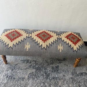 Könnte beinhalten: Rechteckige gepolsterte Bank mit grauem Stoffbezug mit einem geometrischen Muster in Rot, Beige und Marineblau. Die Bank hat vier gedrechselte Holzbeine. Die Bank ist etwa 122 cm lang.