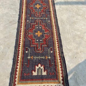 May include: A rectangular, dark blue runner rug with a geometric pattern. The design features red, yellow, and white accents, with a fringed edge. The rug has a central pattern with cross-like shapes. The text "Jaypore Carpet" is visible.