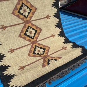 May include: Beige and brown woven rug with geometric diamond and arrow patterns. The rug has a black border with fringe and the text "Jaypore Carpet" in the bottom left corner. The rug is on a blue surface.
