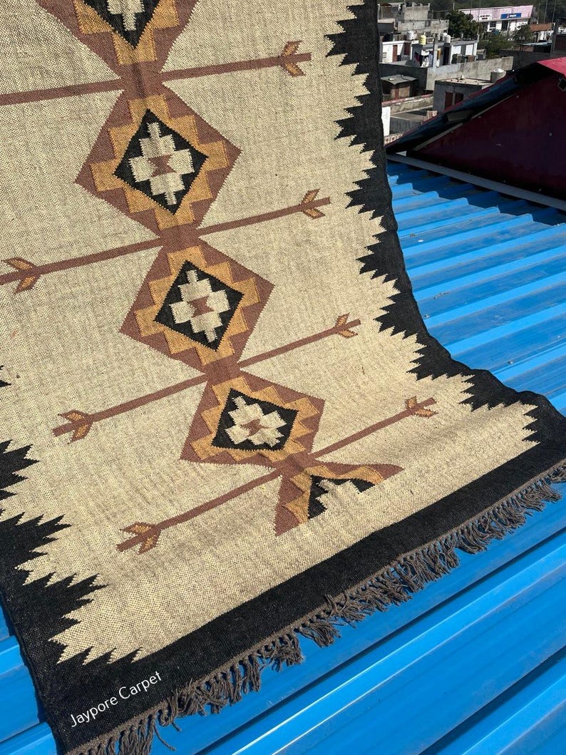 May include: Beige and brown woven rug with geometric diamond and arrow patterns. The rug has a black border with fringe and the text "Jaypore Carpet" in the bottom left corner. The rug is on a blue surface.