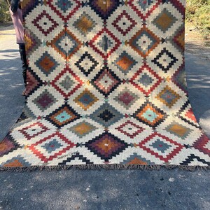 May include: A large, rectangular rug with a geometric diamond pattern in shades of cream, red, blue, orange, and black. The rug has a fringed edge and the text "Jaypore Carpet" is visible. The rug is held up by a person.