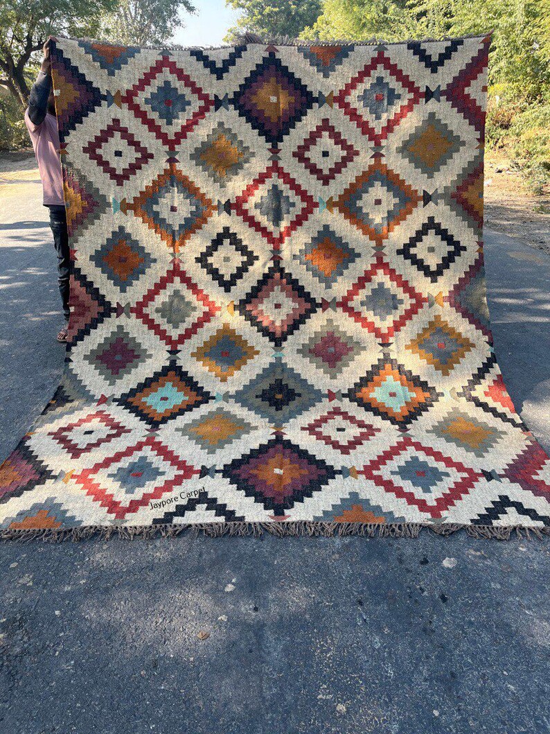 May include: A large, rectangular rug with a geometric diamond pattern in shades of cream, red, blue, orange, and black. The rug has a fringed edge and the text "Jaypore Carpet" is visible. The rug is held up by a person.