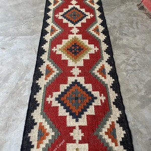 Peut inclure: Un long tapis de couloir rectangulaire présentant un motif géométrique répétitif en forme de losange dans des tons rouge, crème, bleu foncé, orange et vert olive. Les bords sont noirs et le texte Jaypore Carpet est imprimé en blanc.