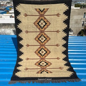 May include: A rectangular beige rug with a geometric pattern in brown and black. The rug features a central design of diamonds and arrows, framed by a black border with a zigzag edge. The text "Jaypore Carpet" is visible at the bottom.