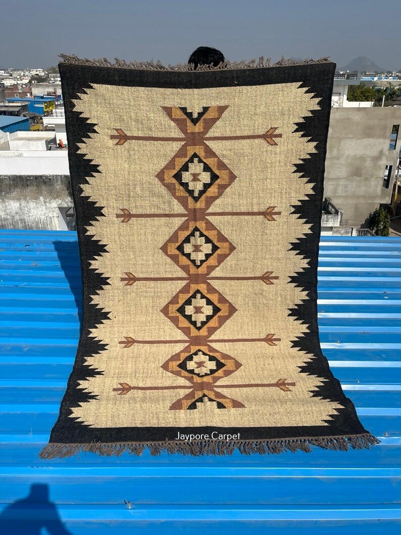 May include: A rectangular beige rug with a geometric pattern in brown and black. The rug features a central design of diamonds and arrows, framed by a black border with a zigzag edge. The text "Jaypore Carpet" is visible at the bottom.