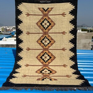 May include: A rectangular beige rug with a geometric pattern of diamonds and arrows in brown and black. The rug has a black border with a zigzag design and fringe at the bottom. The text "Jaypore Carpet" is visible at the top.