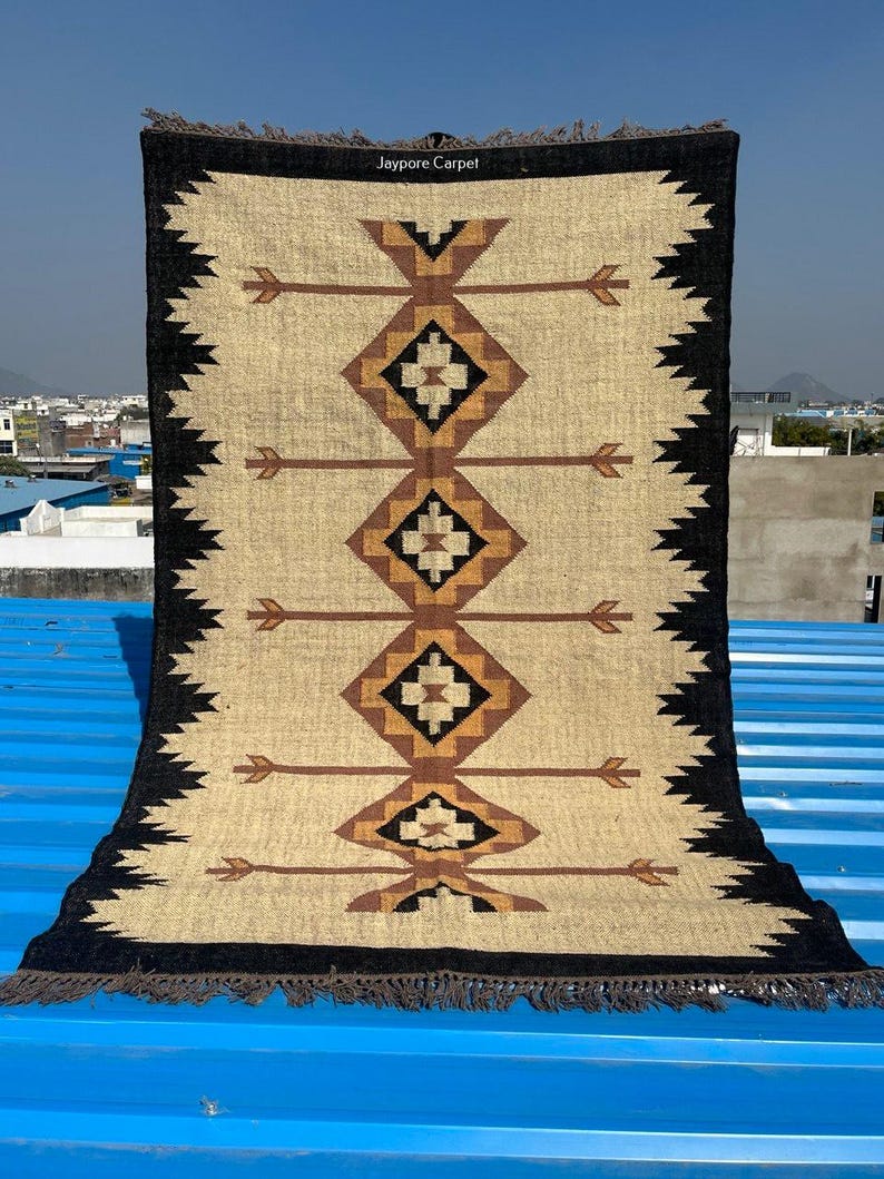 May include: A rectangular beige rug with a geometric pattern of diamonds and arrows in brown and black. The rug has a black border with a zigzag design and fringe at the bottom. The text "Jaypore Carpet" is visible at the top.