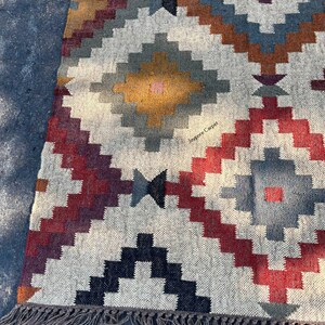 May include: A rectangular, woven rug with a geometric pattern in shades of red, blue, brown, and yellow on a cream background. The rug has a fringe along one edge and the text "Jaypore Carpet" is visible.